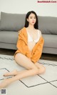 A woman sitting on the floor in a white bra and orange jacket.